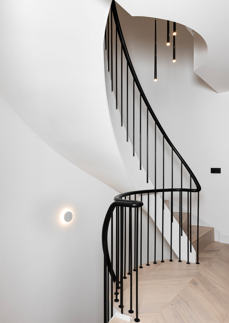 Bespoke luxury helical staircase with black metal spindles and handrail that is lit with minimalist moonlike wall light and slimline overhanging pendant light